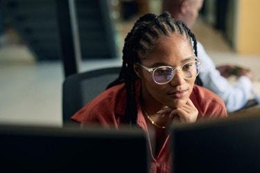 A woman wearing glasses sits in front of two computer monitors. She is analyzing and refining data.