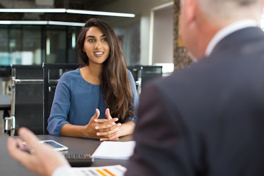 A long-haired young woman sits at a dark gray table in a large open office space. Her hands are folded on the table in front of her, next to an open notebook and a calculator. A white-haired man in a suit sits across from her and gestures with his left hand; he is facing away from the viewer and is out of focus.