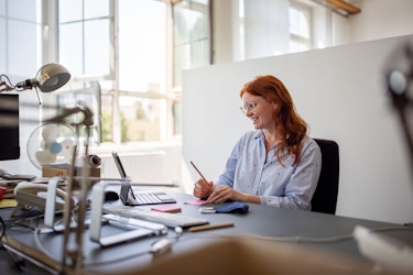 A woman working at her desk is engaged in a video call on her laptop. She is making notes during the teleconference.