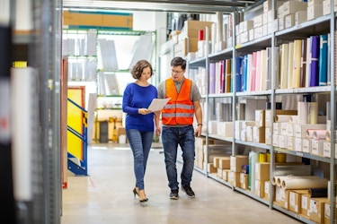 Two employees are walking between rows of shelving units in a warehouse. The person on the left, a woman with bobbed brown hair, is holding a packet of papers. The person on the right, a man wearing glasses and an orange high-visibility vest, looks over at the papers in his coworker's hands. The shelves in the background are filled with cardboard boxes, cardboard tubes, and multicolored folders.