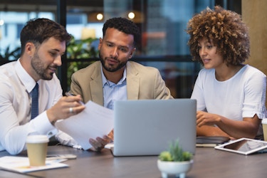 Three people sit around a table. The person on the far left wears a shirt and tie and holds up a piece of paper, which the other two people, a man and a woman, look at with interest. On the table are an open laptop, an electronic tablet, and a takeout coffee cup.
