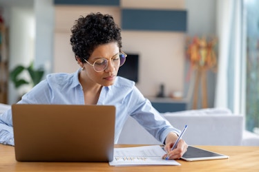 A woman entrepreneurs works at a desk in her home updating her business plan. Her laptop is open, and she is checking figures on a paper before her to ensure they are correct.