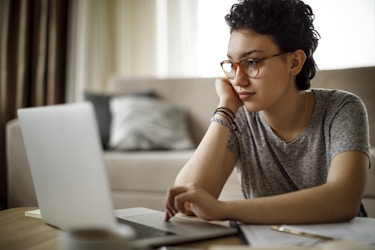 A young woman sits on the floor of a living room, leaning her elbows on a coffee table and propping up her head with one hand. She uses the other hand to operate the track pad on a laptop. The woman has short, dark, curly hair and wears red-rimmed glasses and a gray T-shirt.