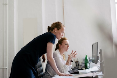  Two women are pictured in an office setting. They are both leaning over a desk looking at  a computer screen. One of the women is illustrating a point by gesturing with her hands. 