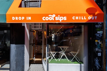 The exterior of a Cool Sips location in New York City. The shop has a glass front under an orange awning. The awning is printed with Cool Sips' logo between the phrases "DROP IN" and "CHILL OUT." Through the glass can be seen a slightly elevated seating area with two chairs and a small table on top of fake grass.