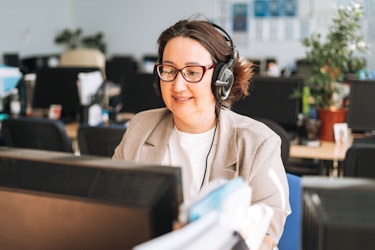  A woman office worker uses a headset while she types notes using a desktop computer. 
