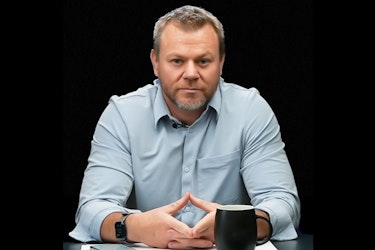A shot of Daniel Ramsey, CEO and Founder of MyOutDesk, sitting at a desk with his hands together. He has graying dark blond hair and a beard, and he's wearing a pale blue button-up shirt and a smart watch.