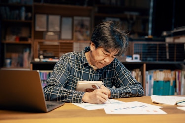  A businessman sits at a table. He is holding a notebook in his left hand. He has papers spread out on the table in front of him and a laptop. He is reviewing financial data. 