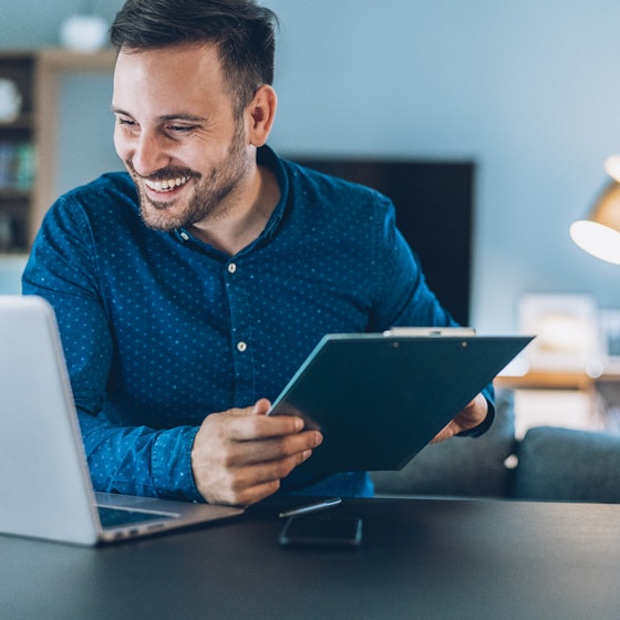 A man working from home sits at a table. He holds an electronic table in his hands, but his smiling face is turned toward his laptop.