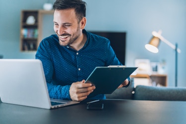 A man working from home sits at a table. He holds an electronic table in his hands, but his smiling face is turned toward his laptop.