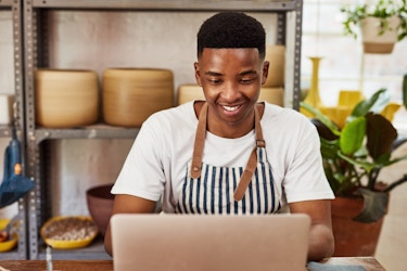 A young man sits at a laptop smiling. He wears a white T-shirt under a blue-and-white striped apron. Behind him are shelving units filled with large ceramic pots.