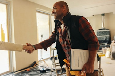 A man wearing a toolbelt and carrying a clipboard shakes a woman's hand. The woman is not pictured in the photo. The man is a general contractor meeting with his client.