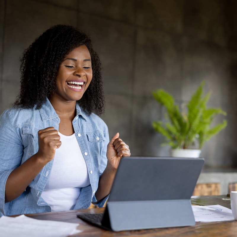 A young woman is sitting at a table in an office. She has a computer tablet propped up in front of her. She is grinning because she just received very good news.
