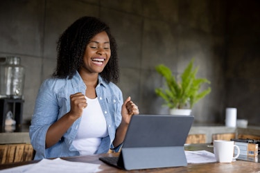 A young woman is sitting at a table in an office. She has a computer tablet propped up in front of her. She is grinning because she just received very good news.