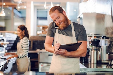 A male small business owner wearing an apron and cradling a phone between his shoulder and cheek writes notes down on a clipboard while he talks on the phone.