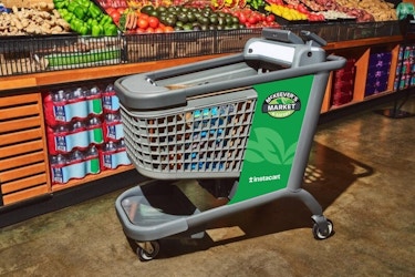A shot of an Instacart Caper Cart, which allow customers to scan and pay for products without waiting in line at checkout, standing in front of a grocery store produce section. The cart is metallic gray with green on the sides. The logo for the grocery store (McKeever's Market and Eatery) and the Instacart logo are also printed on the side of the cart. Where the child's seat would normally be is a touchscreen and credit card scanner..