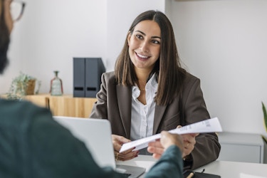 A woman job candidate smiles as she hands her resume to a small business owner. She is interviewing for a position at a small company.