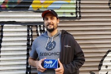  A shot of Dr. Jonathan Scheiman, Co-founder and CEO of FitBiomics, standing in front of a mural and holding a small blue box of FitBiomics' Nella probiotics capsules. Jonathan is a white man with a beard. He's wearing a black-and-red baseball cap and a hooded gray T-shirt with the FitBiomics logo in black on the front. Draped over one shoulder is a black hoodie with FitBiomics's logo on one side. 