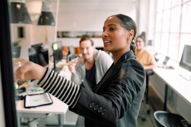 A small group of employees are meeting informally. A woman stands in front of a whiteboard and is pointing at it. The group is discussing a business strategy.