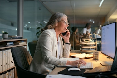 A woman is seated at a desk in a modern office. She is talking on the phone and typing on a desktop computer.