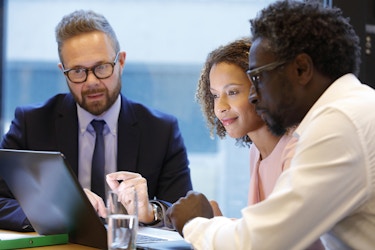Three people sit around a table and look at the screen of an open laptop. The person on the left is a man in a suit and glasses. He is speaking to the couple on the right, a man and woman looking at the screen of the laptop with great interest.