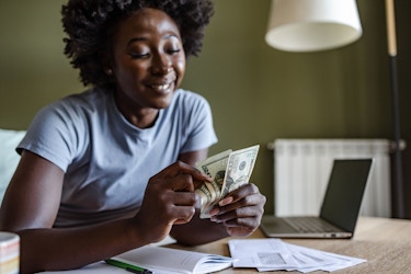 A woman holds up several $20 U.S. bills. She is smiling. She has some letters spread in front of her and a laptop computer.