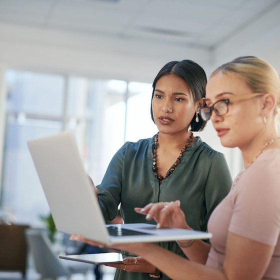Two women business owners hold a laptop computer, looking at its screen, while they discuss an issue.