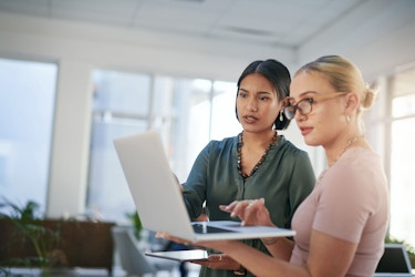 Two women business owners hold a laptop computer, looking at its screen, while they discuss an issue.