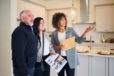 Three individuals stand in a modern white kitchen. A real estate agent is showing a couple the home's interior and highlighting various features of the home.