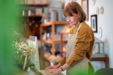 A woman home decor shop owner stands at a desk in her store. She is typing on the keyboard of a desktop computer. She is researching keywords she can use in her local search engine optimization strategy.