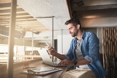 A man sits at a counter in a cafe. He has one hand on the keyboard of an open laptop while his other hand holds up a smartphone. The man has dark hair and a beard starting to grow in. He's wearing a chambray shirt over a gray T-shirt. On the other side of the counter is a room on a lower level lit with golden light streaming in front outside.