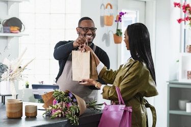 A man in a flower shop hands a paper bag to a female customer. The man wears glasses, a dark blue long-sleeved shirt, and a gray apron. The woman has long dark hair and wears a khaki green overcoat. A bouquet of flowers sits on a table between the sales associate and the customer. In the background, flowers grow from pots hanging from the walls and in shelves around the room.