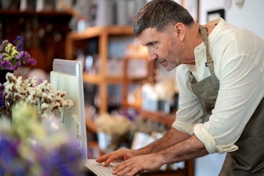The male owner of a home decor store hunches over a desktop computer while he types on the keyboard.