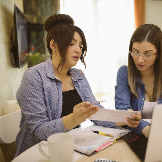 Two women co-business owners are seated at a table. They are reviewing receipts in preparation for their business taxes. One woman holds a paper while another woman types something into her laptop.