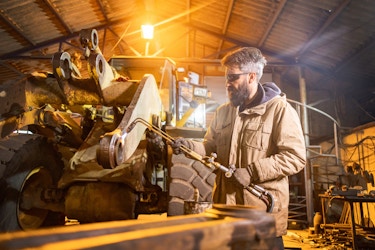 A man in a workshop and uses a blowtorch to weld something on a large construction vehicle. The man has gray hair and a beard, and he's wearing a tan parka, sunglasses, and thick leather gloves.