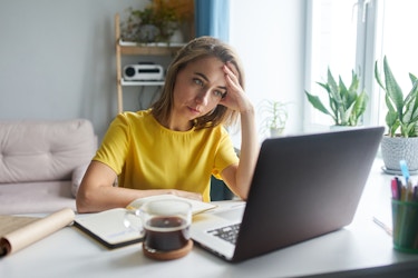 A woman sits at a table in front of an open laptop with her head in one hand and a worn-out look on her face. The woman has shoulder-length blonde hair and she wears a yellow T-shirt.